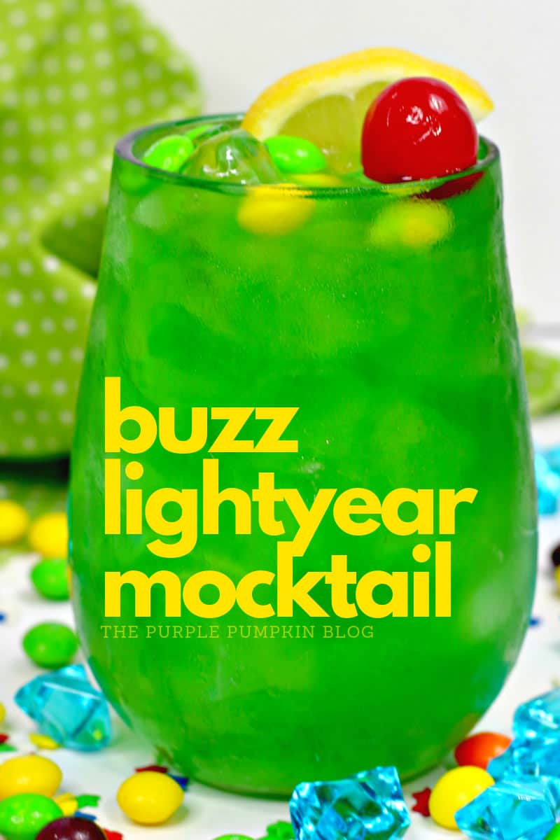 A glass of green drink decorated with a cherry and slice of lemon and green and yellow candies. Sitting on a white table with candies and crystals, and a green and white polka dot napkin in the background.