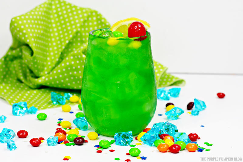 A glass of green drink decorated with a cherry and slice of lemon and green and yellow candies. Sitting on a white table with candies and crystals, and a green and white polka dot napkin in the background.
