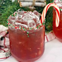 A glass with red, green, and white sprinkles on the rim, filled with red cocktail and ice and garnished with a candy cane.