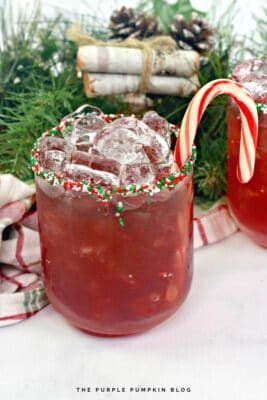 A glass with red, green, and white sprinkles on the rim, filled with red cocktail and ice and garnished with a candy cane.