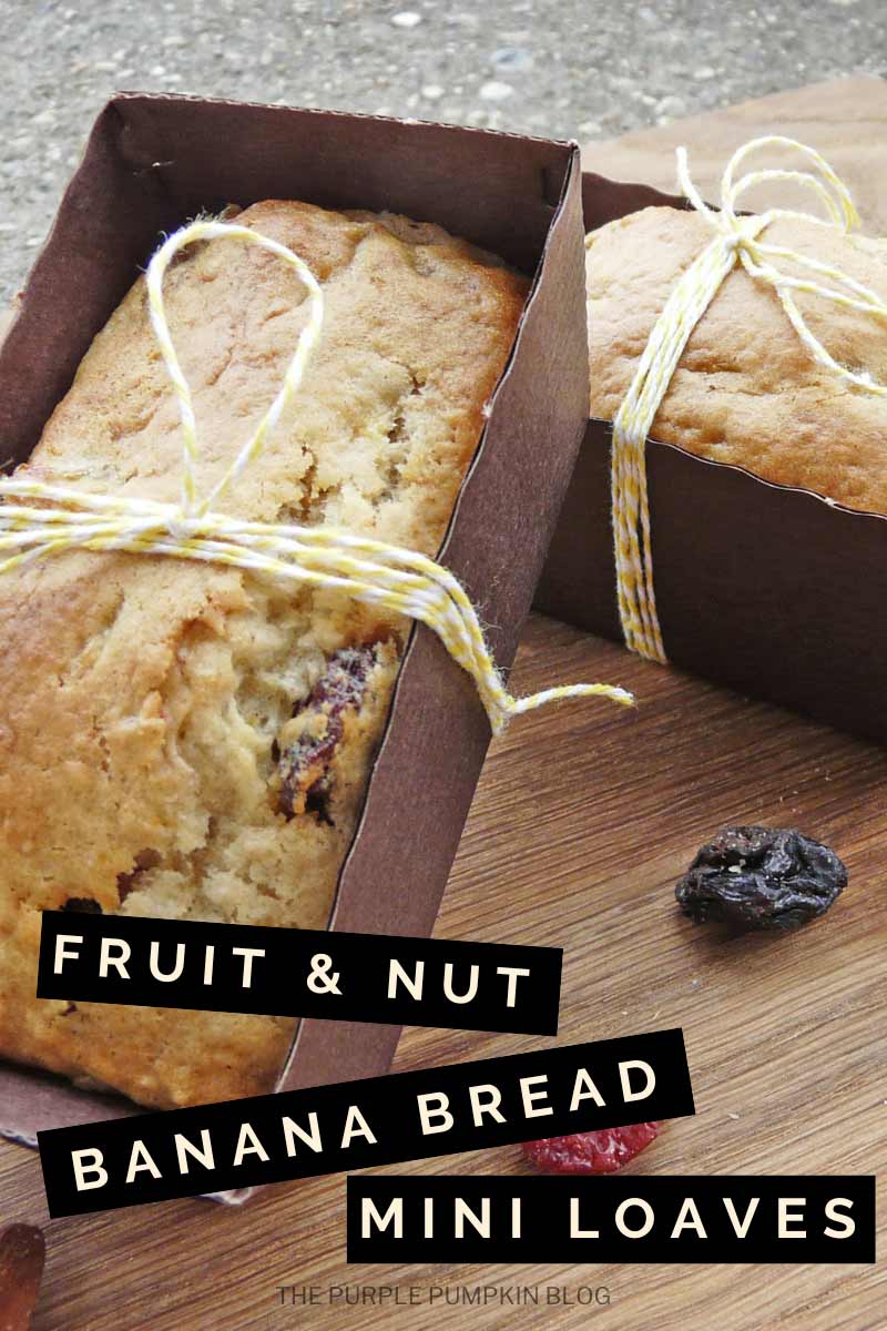 Two mini banana bread loaves, wrapped with bakers twine stacked on a wooden board, with dried fruit scattered around. Text overlay says "Fruit & Nut Banana Bread Mini Loaves"