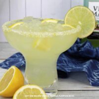 A glass of Italian Margarita filled with crushed ice and garnished with lemon slices and a lime wheel. The drink is rimmed with sugar and served in a classic margarita glass. A bottle of Villa Massa limoncello is blurred in the background.