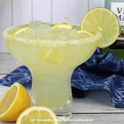 A glass of Italian Margarita filled with crushed ice and garnished with lemon slices and a lime wheel. The drink is rimmed with sugar and served in a classic margarita glass. A bottle of Villa Massa limoncello is blurred in the background.