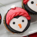 Cupcakes topped with black frosting, that is then decorated with white fondant to create a penguin face, with piped on eyes and beak. A red hat made with fondant sits atop, with holly and snowflake sprinkles. Holiday decorations are in the background.