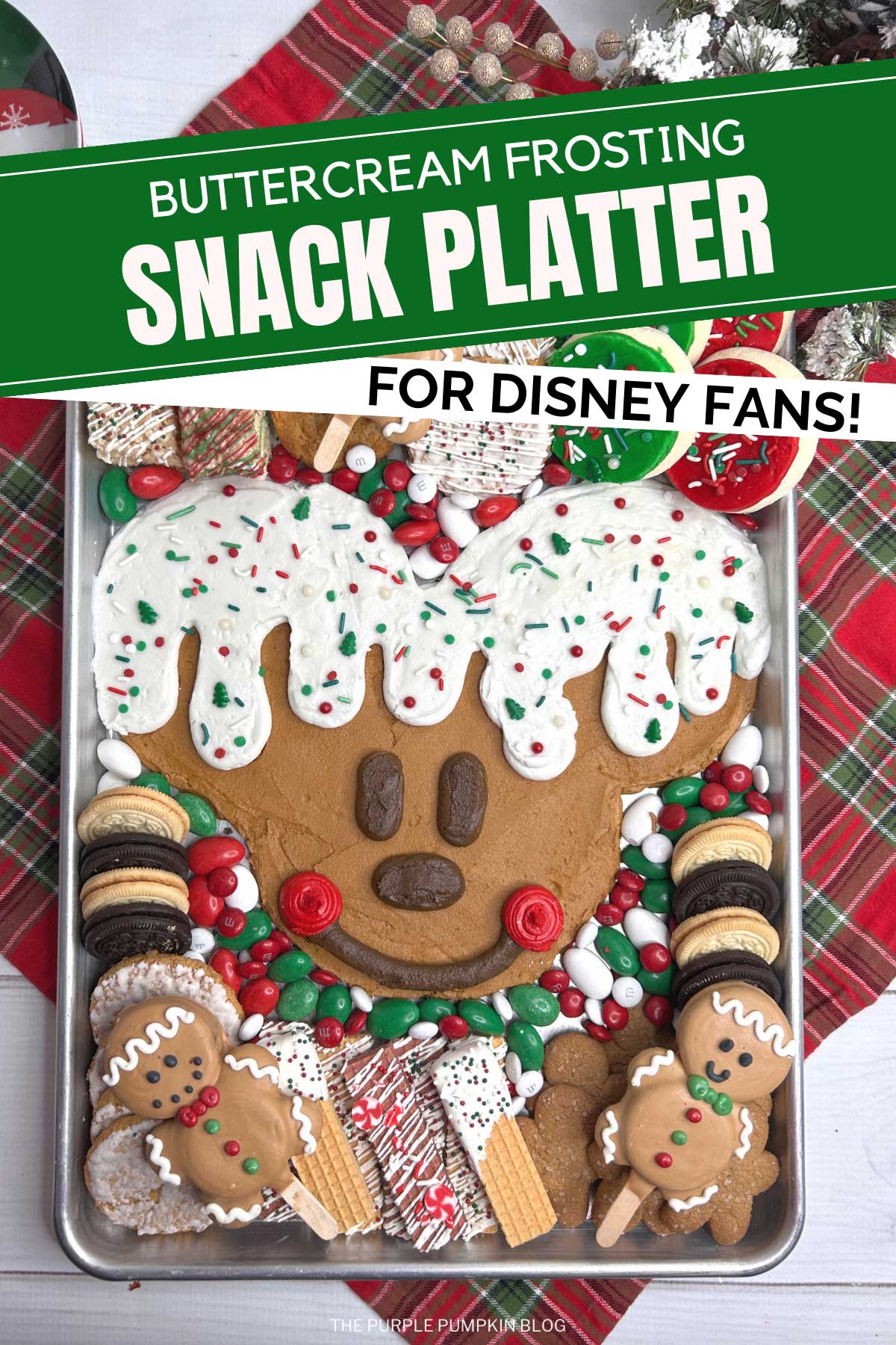A metal tray with Mickey Mouse's head piped on with brown frosting, with white icing dripping down to resemble a gingerbread man. Surrounded by red, white, and green candies, a selection of cookies and other festive treats. The tray is surrounded by festive decorations and tableware.
