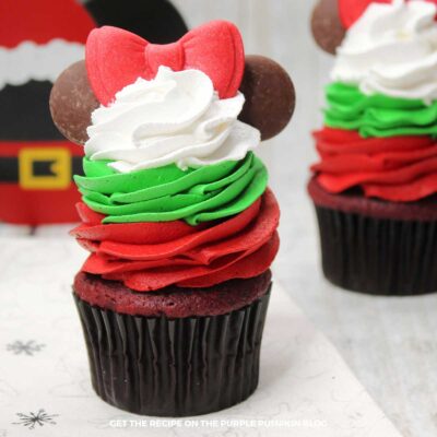 Red velvet cupcakes topped with a swirl of red frosting, topped with a swirl of green frosting, topped with a swirl of white frosting. A red fondant bow sits on top of the stack of tricolor frosting, with 2 round chocolate wafers either side to resemble Minnie Mouse's ears.