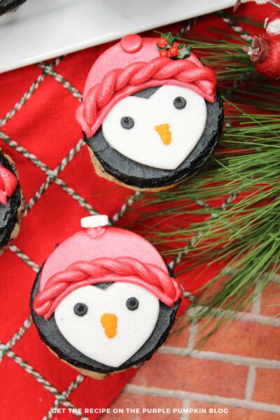 Cupcakes topped with black frosting, that is then decorated with white fondant to create a penguin face, with piped on eyes and beak. A red hat made with fondant sits atop, with holly and snowflake sprinkles. Holiday decorations are in the background.
