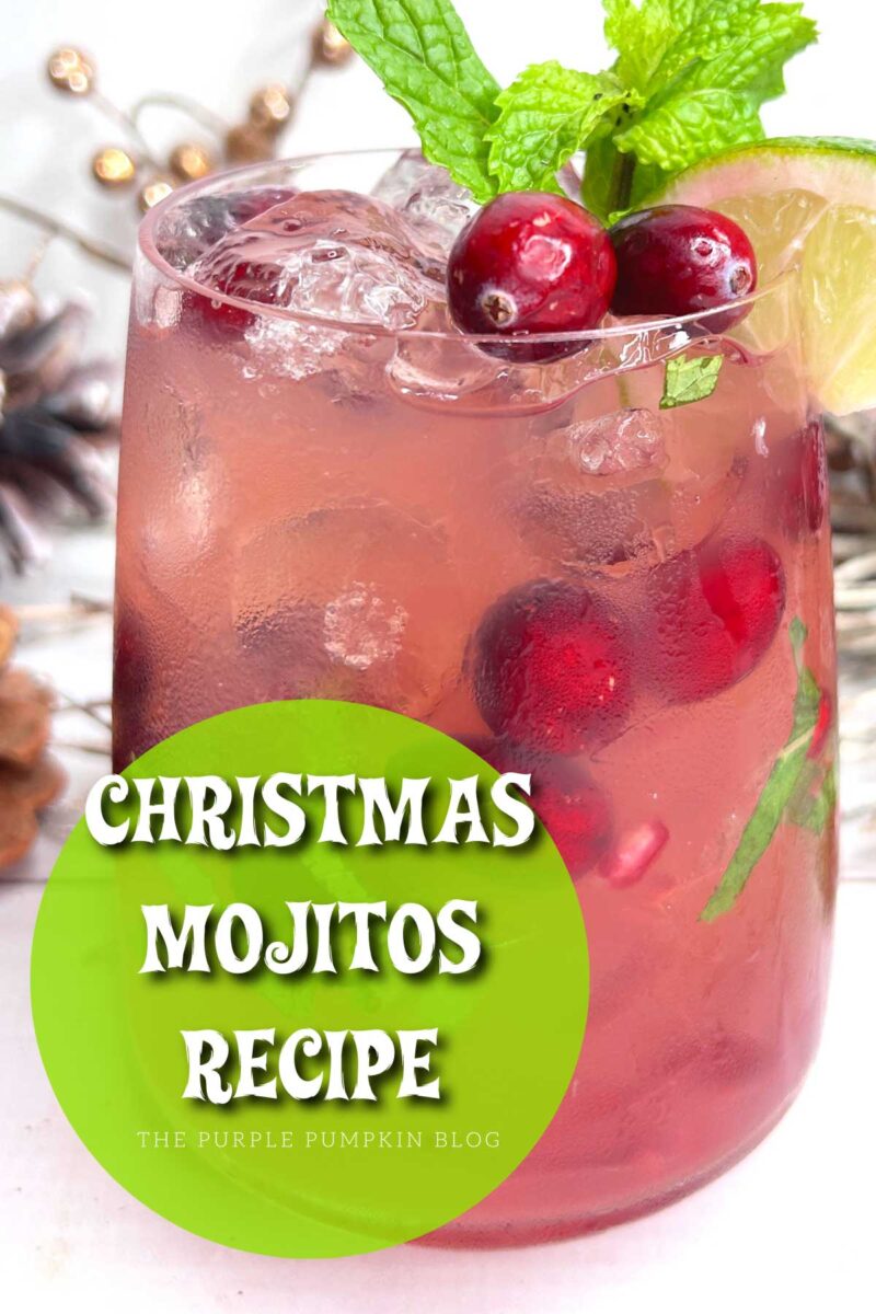 A cocktail glass filled with red liquid, ice, chopped fresh mint leaves, and fresh cranberries. A sprig of fresh mint, a couple of cranberries, and a wedge of lime garnish the glass. In the background are festive decorations and a cocktail shaker.