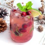 A cocktail glass filled with red liquid, ice, chopped fresh mint leaves, and fresh cranberries. A sprig of fresh mint, a couple of cranberries, and a wedge of lime garnish the glass. In the background are festive decorations and a cocktail shaker.