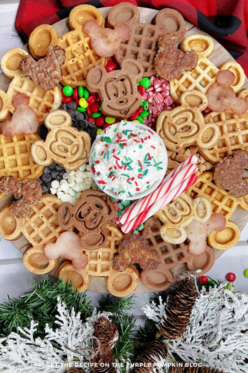 A round board topped with 2 kinds of Mickey Mouse waffles, Mickey-shaped sausage patties and ham, a bowl of whipped cream, candy canes, and a variety of holiday candies. Festive decorations surround to board.