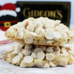 A stack of cookies loaded with white chocolate chips and coarse sugar sprinkles. In the background is a Mickey Mouse Santa ornament and a box from Gideon's Bakehouse at Disney Springs.