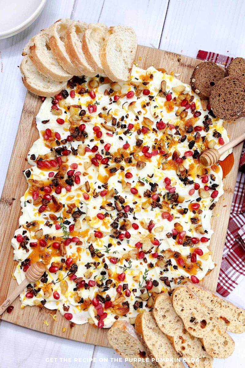 Wooden board covered in a layer of butter topped with orange zest, pomegranate seeds, fresh herbs, roasted garlic, and salt. Some slices of bread are laid next to the board.