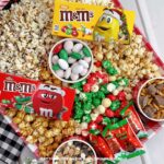 A red gingham tray, filled with pots of candy, pretzels, and chocolate chips, surrounded by various popcorn flavors, and topped with boxes of M&Ms and packets of Reese's Christmas Trees.