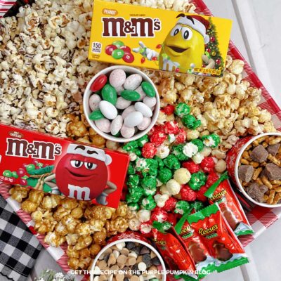 A red gingham tray, filled with pots of candy, pretzels, and chocolate chips, surrounded by various popcorn flavors, and topped with boxes of M&Ms and packets of Reese's Christmas Trees.