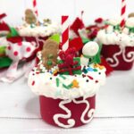 A giant marshmallow, dipped in red candy melts with a red-and-white striped straw inserted. The marshmallow is decorated with melted white candy to resemble snow, and topped with festive sprinkles, and a gingerbread man and Christmas gift made from melted candy. More decorated marshmallows are in the background.