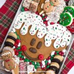 A metal tray with Mickey Mouse's head piped on with brown frosting, with white icing dripping down to resemble a gingerbread man. Surrounded by red, white, and green candies, a selection of cookies and other festive treats. The tray is surrounded by festive decorations and tableware.