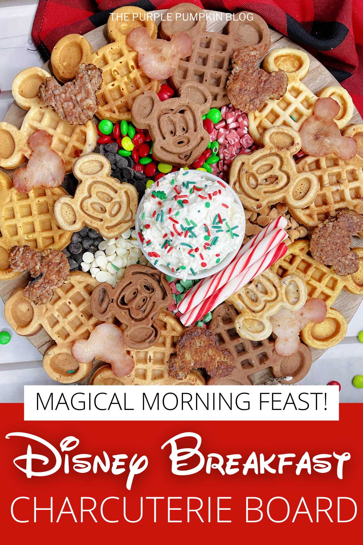 A round board topped with 2 kinds of Mickey Mouse waffles, Mickey-shaped sausage patties and ham, a bowl of whipped cream, candy canes, and a variety of holiday candies. Festive decorations surround to board.