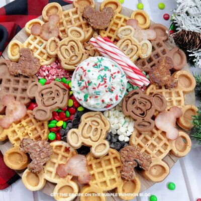A round board topped with 2 kinds of Mickey Mouse waffles, Mickey-shaped sausage patties and ham, a bowl of whipped cream, candy canes, and a variety of holiday candies. Festive decorations surround to board.