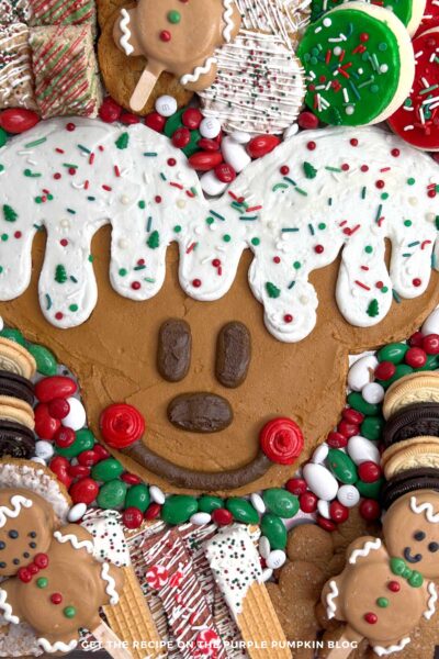 A metal tray with Mickey Mouse's head piped on with brown frosting, with white icing dripping down to resemble a gingerbread man. Surrounded by red, white, and green candies, a selection of cookies and other festive treats. The tray is surrounded by festive decorations and tableware.