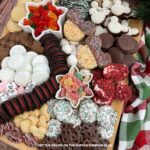 A wooden board covered with a variety of Mickey Mouse sweet treats - cookies, chocolate-covered pretzels, gummies, coconut patties, and rice krispy treats. Additional generic holiday treats and cookies are also on the board. The board is sat on a festive cloth, with a stack of Disney plates to the side.