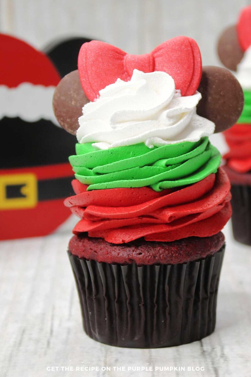 Red velvet cupcakes topped with a swirl of red frosting, topped with a swirl of green frosting, topped with a swirl of white frosting. A red fondant bow sits on top of the stack of tricolor frosting, with 2 round chocolate wafers either side to resemble Minnie Mouse's ears.