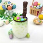 Glass of white cocktail with chocolate rim, Easter Bunny candy, toasted green coconut and mini eggs on top, surrounded by Easter decorations