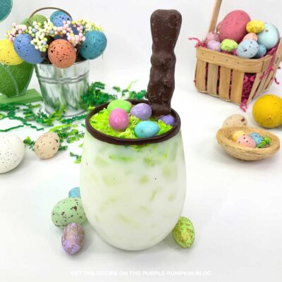 Glass of white cocktail with chocolate rim, Easter Bunny candy, toasted green coconut and mini eggs on top, surrounded by Easter decorations
