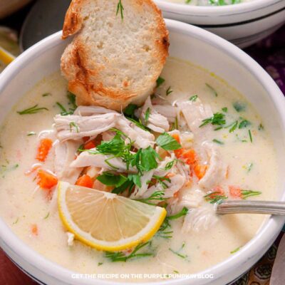 Bowl of creamy chicken soup, topped with fresh herbs, lemon slice, and toasted piece of bread. Other ingredients surround.