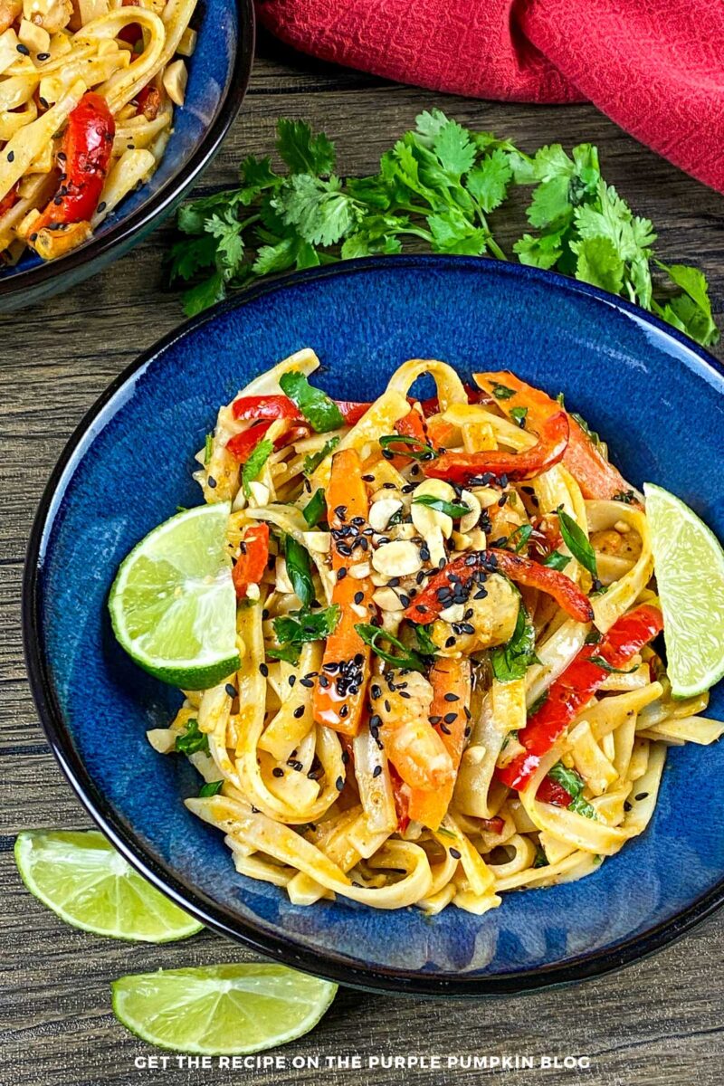 Bowl of noodles, shrimp and veggies in a creamy sauce, garnished with peanuts, sesame seeds and lime wedges.