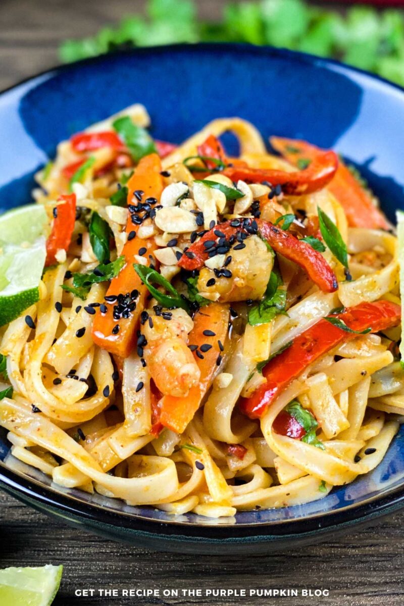 Bowl of noodles, shrimp and veggies in a creamy sauce, garnished with peanuts, sesame seeds and lime wedges.