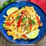 Bowl of noodles, shrimp and veggies in a creamy sauce, garnished with peanuts, sesame seeds and lime wedges.