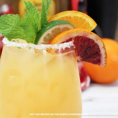 Glass of orange cocktail with slices of citrus fruit and mint leaves as garnish and a salted rim. Bottles of alcohol are in the background.