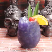 Glass filled with deep purple cocktail, garnished with a cocktail cherry, slice of orange, and pineapple leaves. Skull candle holders are in the background.