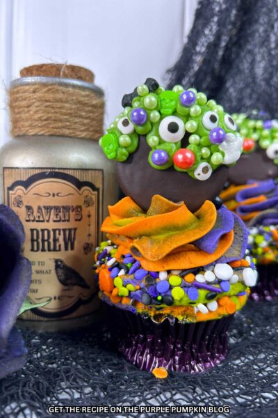 Cupcakes topped with multi-colored frosting (orange, blue, and green) and sprinkles and an Oreo dipped in chocolate and covered in green icing and a variety of sprinkles including eyeballs, bats, and ghosts to resemble a bubbling cauldron. Halloween decor surrounds the cupcakes.