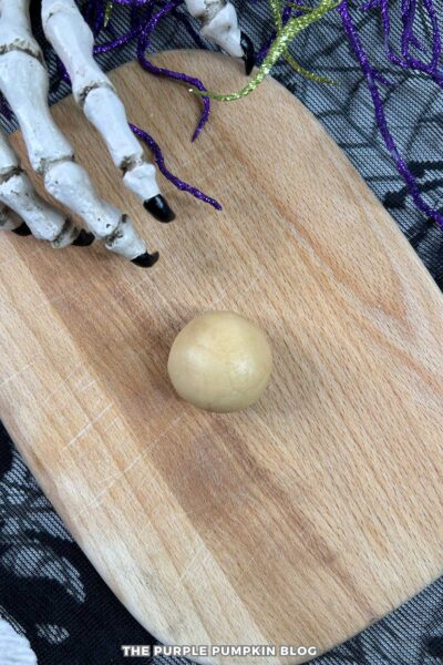 Round ball of cake mixture placed on a wooden board, ready to be coated in candy melts. A decorative skeleton hand and sparkly purple and green garland add a spooky Halloween touch.