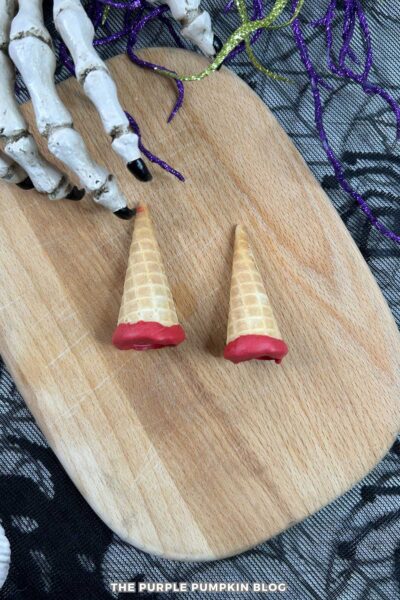 Two small waffle cone pieces dipped in red candy melts, placed on a wooden board beside a skeleton hand and purple garland for Halloween decoration.