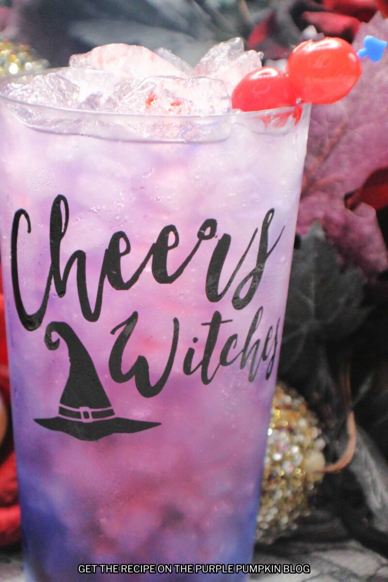 A glasses embossed with "Cheers Witches" and a witch hat, filled with a pink-purple drink and garnished with cocktail cherries. Halloween decorations are in the background.