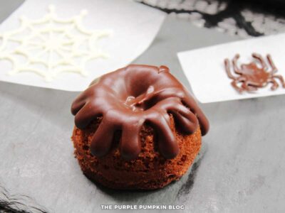 Mini chocolate bundt cake with chocolate ganache dripped on top. A white chocolate web and chocolate spider piped on wax paper is in the background.