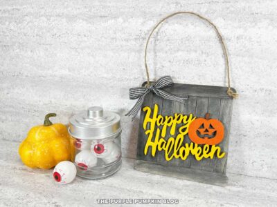 A sign made with craft sticks and decorated with the words Happy Halloween and Jack-o'lantern and a ribbon bow. A jar of fake eyeballs and mini pumpkin are sat beside it.