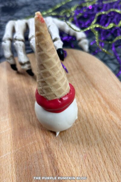 Partially assembled Halloween cake pop made from a white candy-coated cake ball topped with a waffle cone dipped in red candy melts. Displayed on a wooden board with a decorative skeleton hand and purple garland in the background.