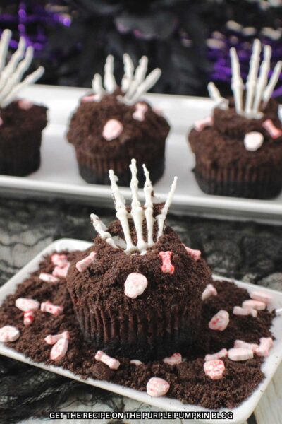 Mini chocolate cheesecakes, covered with crumbled cookies to resemble dirt, with a fondant skeletal hand poking out of the top and skull and bone shaped sprinkles. Halloween decor is in the background.