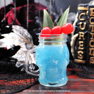 Skull-shaped glass mug filled with blue cocktail and garnished with red cherries and green leaves. Halloween decor is in the background.