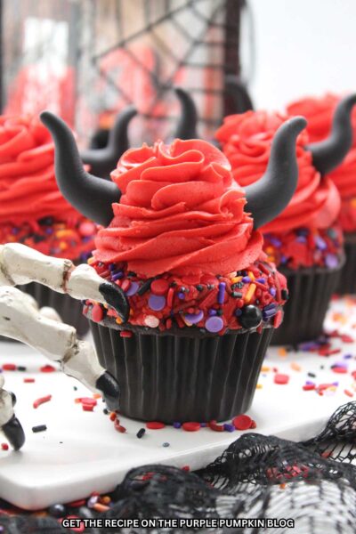 Cupcakes topped with bright red frosting and sprinkles with a swirl of frosting on top with black horns poking out. Halloween decor in the background.