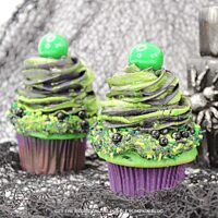 Cupcake topped with frosting and green and black sprinkles, with another swirl of black and green frosting on top with a green candy ball to finish. Halloween decor is in the background.