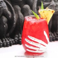 Glasses with a skeleton hand design filled with red cocktail, and garnished with a cherry, pineapple wedge and leaves.