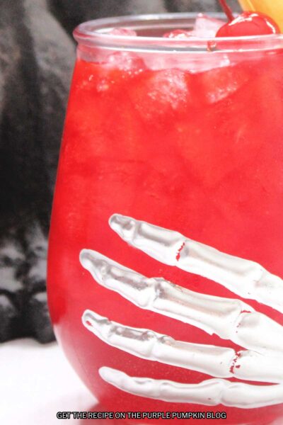Glasses with a skeleton hand design filled with red cocktail, and garnished with a cherry, pineapple wedge and leaves.
