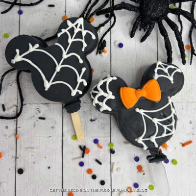 Mickey Mouse-shaped Oreo Cookie Pops, covered with black candy melts and white candy spiderwebs. Minnie has an orange bow.with Halloween decorations scattered around.