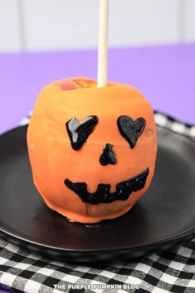An apple dipped in orange candy melts and decorated with black icing to create a Jack-o'-Lantern face.