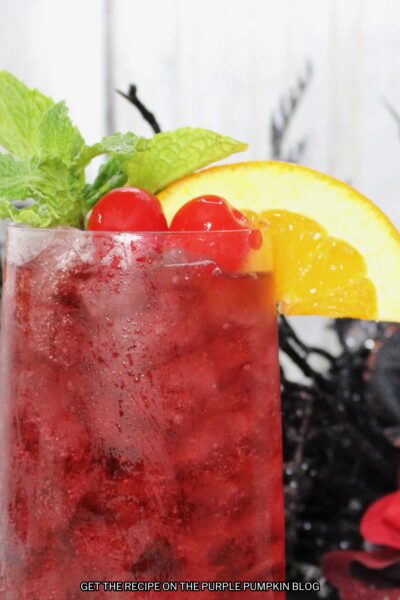A glass of deep red cocktail garnished with cocktail cherries, slice of orange, and mint leaves. Halloween decorations are in the background.