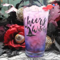 A glasses embossed with "Cheers Witches" and a witch hat, filled with a pink-purple drink and garnished with cocktail cherries. Halloween decorations are in the background.